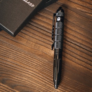 May include: Black tactical pen with a textured grip and a pocket clip, resting on a wooden surface. The pen has a pointed tip and a logo on the top. A black box is partially visible in the background.