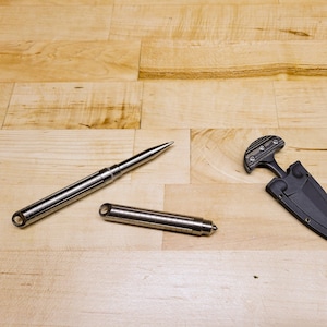 May include: Two silver metal self-defense tools on a light wood surface. One tool is a pen-shaped device with a pointed tip. The other tool is a small, black, folding knife.