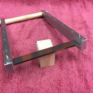 May include: A metal frame with a wooden dowel and a metal blade resting on a wooden block. The frame has multiple holes along the sides.