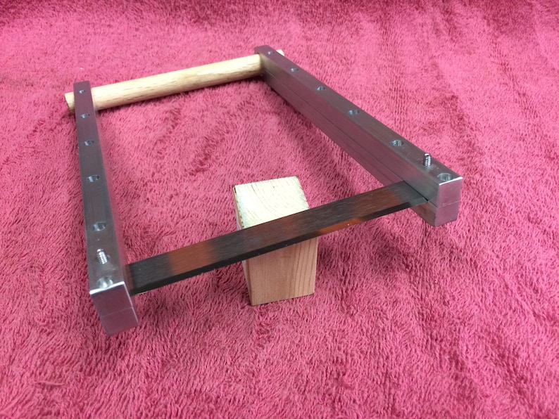 May include: A metal frame with a wooden dowel and a metal blade resting on a wooden block. The frame has multiple holes along the sides.