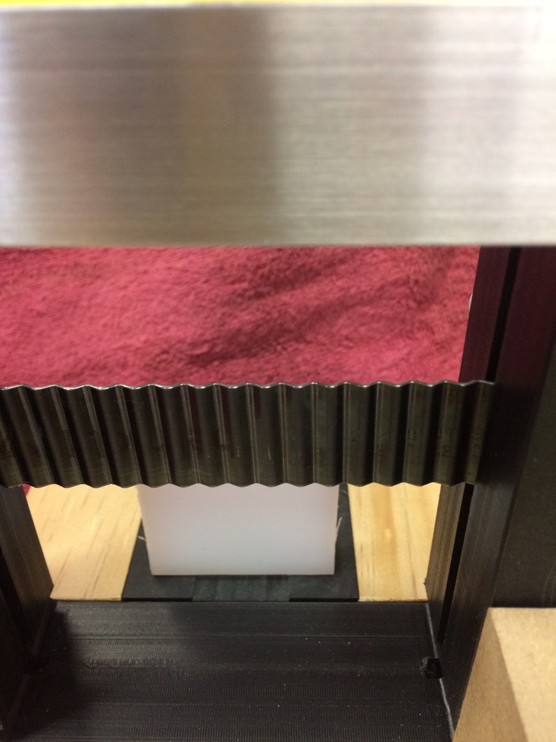 K&ouml;nnte beinhalten: Nahaufnahme eines schwarzen Metallrahmens mit einem wei&szlig;en rechteckigen Objekt darin. Der Rahmen hat oben eine Wellblechleiste und unten eine schwarze Gummileiste. Das Objekt liegt auf einer Holzoberfl&auml;che.