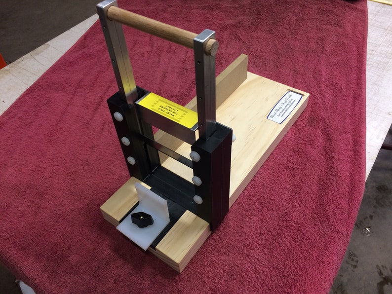 May include: A wooden sausage stuffer with a metal frame and a handle. The stuffer has a black plastic base and a white plastic knob. The stuffer is sitting on a red towel.