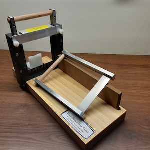 Peut inclure: Un coupe-savon en bois avec un cadre en métal et une poignée en bois. Le coupe-savon est sur une table en bois. Le texte sur l'étiquette indique "Wooden Body & Soap Cutter,  Phone Number: (225) 479-3973,  et l'adresse e-mail est incluse."