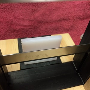 May include: A white rectangular block sits on a wooden surface, partially obscured by a black metal frame. The frame has a silver metal bar across the top.