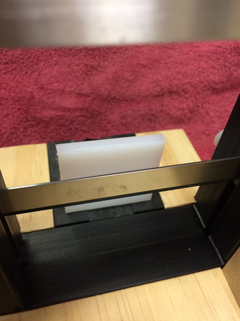May include: A white rectangular block sits on a wooden surface, partially obscured by a black metal frame. The frame has a silver metal bar across the top.