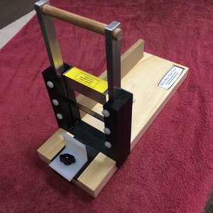 Può includere: Uno stuffer per salsiccia in legno con un telaio in metallo e una maniglia. Lo stuffer ha una base in plastica nera e una manopola in plastica bianca. Lo stuffer è seduto su un asciugamano rosso.