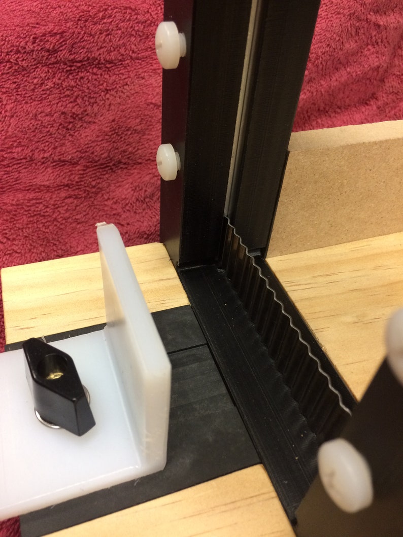 K&ouml;nnte beinhalten: Nahaufnahme eines schwarz-wei&szlig;en 3D-gedruckten Teils mit einer wei&szlig;en Kunststoffhalterung, die daran befestigt ist. Das Teil hat eine gezahnte Kante und ist auf einer Holzoberfl&auml;che montiert.