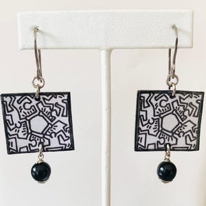 Pu&ograve; includere: Un paio di orecchini quadrati con un design astratto in bianco e nero. Ogni orecchino ha un bordo nero, un gancio argentato e una piccola perla nera. Gli orecchini sono esposti su un supporto bianco.
