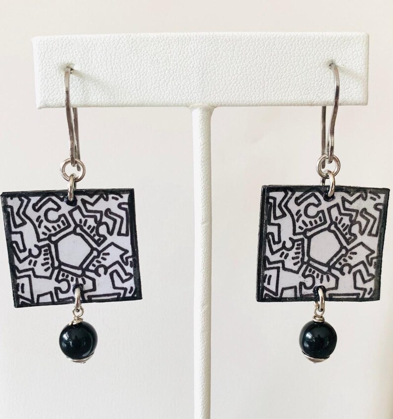 Pu&ograve; includere: Un paio di orecchini quadrati con un design astratto in bianco e nero. Ogni orecchino ha un bordo nero, un gancio argentato e una piccola perla nera. Gli orecchini sono esposti su un supporto bianco.