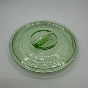 May include: A round, light green glass lid with a central handle. The lid has a slightly raised rim and a clear, smooth surface. The handle is oval-shaped and has a raised, curved design. The glass has a slight transparency.