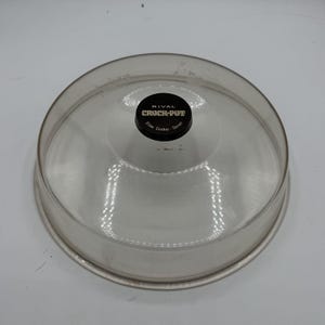 May include: A round, clear glass lid for a slow cooker with a black circular knob in the center. The knob features the white text RIVAL CROCK-POT Slow Cooker - Serve. The lid has a slightly domed shape and a smooth, transparent finish throughout.