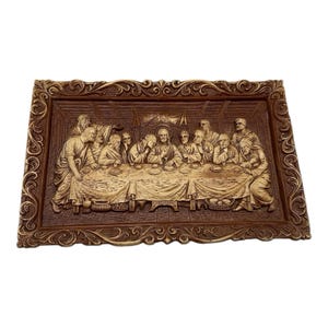 May include: A brown rectangular relief carving depicting the Last Supper. The artwork features detailed figures seated around a table, with a decorative frame. The relief is in shades of brown and beige.