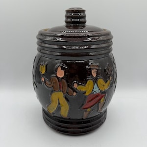 May include: A dark brown ceramic jar with a lid. The jar features a relief design of dancing figures in colorful clothing. The jar is likely used for storage and has a rustic, handcrafted appearance.