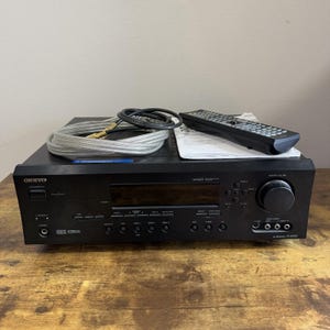 May include: Black Onkyo receiver with a remote control, cables, and a manual. The receiver has a volume knob, multiple input jacks, and a display screen. The remote is black with numerous buttons.