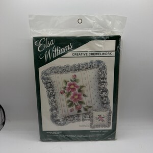 May include: Elsa Williams Creative Crewelwork kit titled Roses and Lace II. The package features a square pillow with a pink rose floral design, white lace trim, and a 12 inch by 12 inch finished size. Includes embroidery yarn, backing, lace, needle, and instructions.