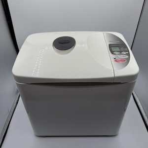 May include: White bread maker with a digital display and control panel. The bread machine has a rectangular shape with a rounded top and a dark gray circular button. The control panel includes buttons for settings and a timer.