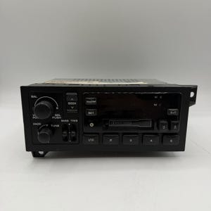 May include: Black car stereo with buttons and knobs for volume, seek, and tuning. Features AM/FM, a digital display, and preset buttons. The stereo has a cassette player and is designed to fit in a car.