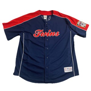 May include: A navy blue baseball jersey with red sleeves and shoulders. The word "Twins" is written in red script across the chest. White piping outlines the front and sides. A patch is on the right sleeve.