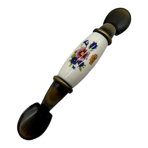 May include: A white ceramic cabinet pull with a floral design in shades of pink, blue, yellow, and green. The pull has a brass finish and is approximately 3 inches long.