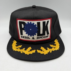 May include: Black baseball cap with a flat brim. The front features a rectangular patch with the word "POLK", a blue gear, and the text "DIESEL & MACHINE". The brim has yellow embroidered leaf designs.