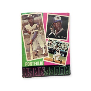 May include: A pink and green baseball card portfolio with the text "Portfolio Hank Aaron" on the front. The portfolio features three baseball players in action.