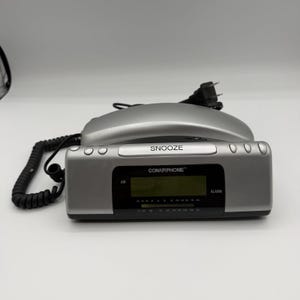 May include: A silver ConairPhone alarm clock with a digital display showing AM radio frequencies. The clock has a snooze button and a coiled cord. The power cord is plugged in.