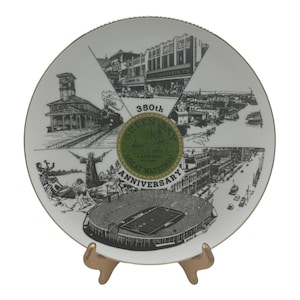 May include: A decorative white plate commemorating the 350th anniversary of Green Bay. The plate features black and white illustrations of historical landmarks, a stadium, and a green and gold emblem with text.