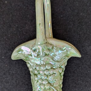 May include: A light green ceramic basket with a handle, featuring a grape cluster design. The basket has a glossy finish with iridescent highlights. The handle arches gracefully above the basket's body.