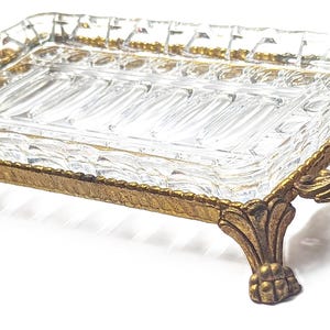 May include: A rectangular clear glass tray with a faceted edge design, set within a gold-toned metal frame. The frame features four ornate, textured legs and two decorative handles on the sides, providing a raised base for the glass insert.
