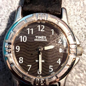May include: A Timex Indiglo wristwatch with a black face and silver bezel. The watch features white numerals and hands, with a black leather band. The watch face has a wave pattern and the Timex Indiglo logo.