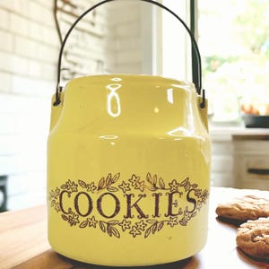 May include: A yellow ceramic cookie jar with a black handle. The word "COOKIES" is written in brown script and surrounded by floral designs. The jar has a milk can shape.