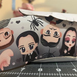 May include: A roll of gray ribbon featuring cartoon illustrations of characters, spiders, and hearts. The ribbon has a textured surface and is partially rolled, revealing the printed design. The characters are in black and white.