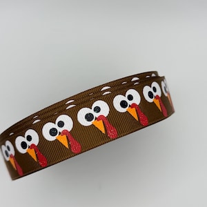 May include: A brown grosgrain ribbon featuring a repeating pattern of cartoon turkey faces. Each turkey has large white eyes with black pupils, a bright orange beak, and a textured red wattle. The ribbon is coiled into a circular shape.
