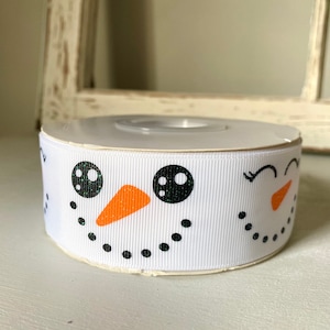 May include: White ribbon with a snowman face design. The snowman has black eyes, a black mouth, and an orange nose.