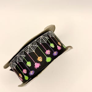 May include: A spool of black grosgrain ribbon featuring a repeating pattern of white spiderwebs at the top edge with colorful cartoon spiders in pink, green, and orange hanging down from thin lines on the black background of the ribbon.