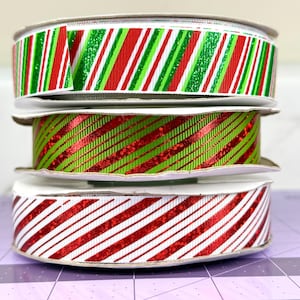 May include: Three spools of decorative ribbon stacked on top of each other. The top spool has green, red, and white striped ribbon. The middle spool has green and red diagonal striped ribbon. The bottom spool has red and white striped ribbon.