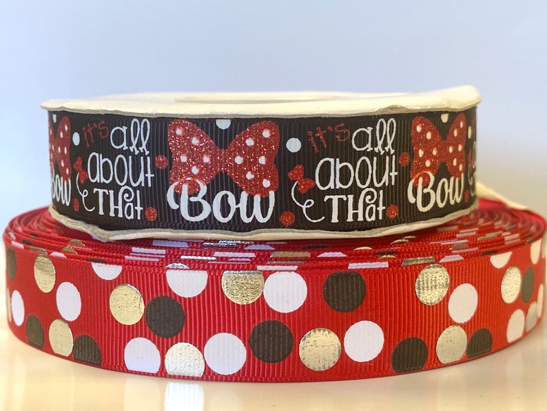 May include: Two spools of ribbon. The top spool is black with the text "it's all about THAT Bow" and a red bow design. The bottom spool is red with gold, white, and brown polka dots. The ribbons are on a white surface.