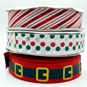May include: Three stacked spools of grosgrain ribbon. The top spool features red glitter diagonal stripes on a white background. The middle spool has a white background with alternating red, green, and silver metallic polka dots. The bottom spool is red with a dark green horizontal stripe and gold glitter belt buckle patterns.