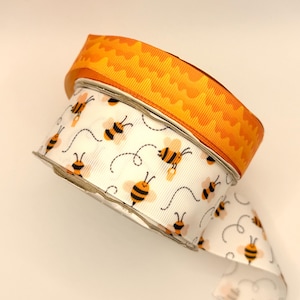 May include: A spool of grosgrain ribbon featuring two distinct designs. The top layer is a solid orange ribbon with a wavy pattern, while the bottom layer is a white ribbon decorated with a repeating pattern of small yellow and black bees.