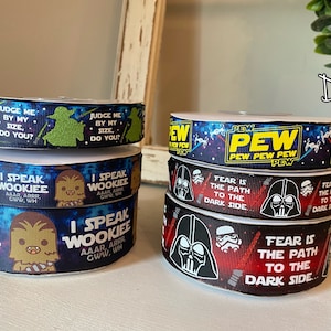May include: A stack of four blue and black ribbon rolls with Star Wars themed designs. The top roll features the text "PEW PEW PEW PEW PEW PEW" in yellow letters. The second roll features the text "FEAR IS THE PATH TO THE DARK SIDE" in white letters. The third roll features a cartoon image of Darth Vader. The bottom roll features the text "I SPEAK WOOKIEE" in white letters.