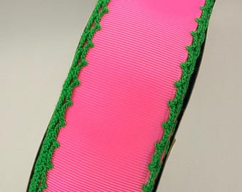 May include: A roll of bright pink grosgrain ribbon featuring a textured, ribbed surface. The edges of the ribbon are finished with a decorative, scalloped green crochet trim that runs along both sides of the length of the pink fabric material.
