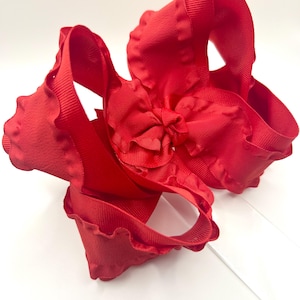May include: A large, vibrant red hair bow. The bow is made of a textured ribbon with ruffled edges and a smaller bow in the center. The bow is a bright, solid red color and is likely for a child or a person who enjoys wearing bows.