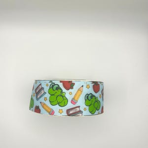 May include: A roll of light blue ribbon featuring a repeating pattern of green cartoon frogs, red apples, yellow pencils, and books. The ribbon has a metallic gold edge and is suitable for crafting and gift wrapping.