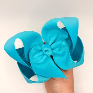 May include: A large, bright turquoise hair bow. The bow is made of grosgrain ribbon and features multiple loops and a knotted center. The bow is held against a plain white background.