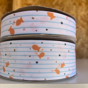 May include: Two spools of white ribbon with a blue and white striped pattern. The ribbon is decorated with orange fish and black stars.
