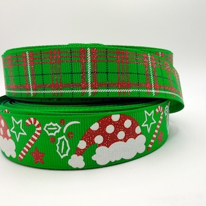 May include: A spool of green ribbon featuring two distinct patterns. The top layer displays a red and white plaid design with glitter accents. The bottom layer shows a green background with white and red Santa hats, candy canes, and stars.