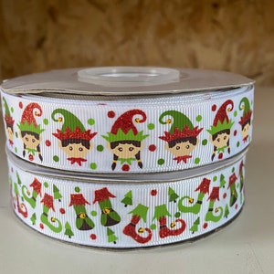May include: Two spools of white ribbon with a repeating pattern of elf faces, green trees, and elf shoes. The elves have red glitter hats and green and red accents. The ribbon is perfect for holiday crafts and gift wrapping.