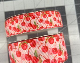 May include: Two rolls of white ribbon with a red cherry print. The cherries are scattered across the ribbon and have green stems and leaves.