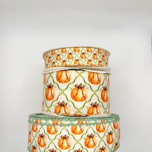 May include: Three stacked spools of white ribbon featuring a repeating pattern of orange pumpkins and green lattice lines. The bottom spool has a decorative scalloped green edge, while the top two spools have smooth, straight edges.