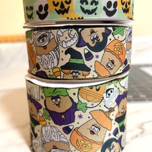 May include: Three rolls of Halloween-themed ribbon. The top roll has a green and yellow background with black jack-o'-lantern faces. The middle roll has a cream background with cartoon characters dressed in Halloween costumes. The bottom roll has a cream background with cartoon characters dressed in Halloween costumes.